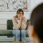 A woman sits on a sofa visibly distressed during a therapy session, hands clasped.